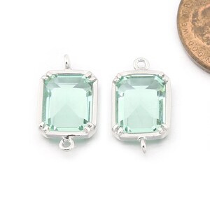 Può includere: Due ciondoli pendenti argentati con pietre di vetro quadrate, trasparenti e di colore verde chiaro. Ogni ciondolo ha un anello nella parte superiore per attaccarlo a una collana o un braccialetto.