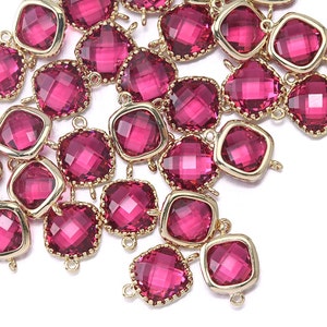 Pode incluir: Um grupo de contas de vidro facetadas cor-de-rosa com conectores de metal dourado. As contas são quadradas e têm uma borda chanfrada.