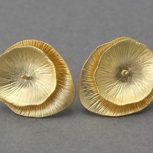 Puede incluir: Un par de pendientes dorados. Cada pendiente presenta un diseño en capas, similar a un pétalo, con una superficie texturizada. Los pendientes tienen cierre de poste y un acabado mate, sobre un fondo gris.