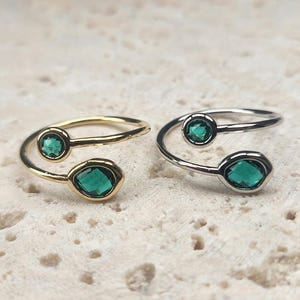 May include: Two open-style rings, one gold-toned and one silver-toned, each featuring two emerald-green gemstones. The gemstones are round and oval-cut. The rings are displayed on a textured, off-white surface.