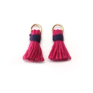 Könnte beinhalten: Zwei rosa Garnquasten mit einem dunkelblauen Band um die Oberseite, befestigt an goldenen Sprungringen.