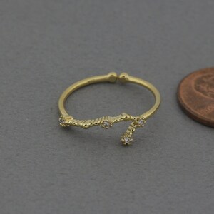 May include: A gold-colored adjustable ring with a constellation design. The ring features small, clear gemstones and is shown on a gray background. The ring is next to a penny for size comparison.