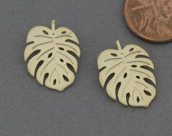May include: Two gold-colored metal charms shaped like monstera leaves. The leaves have a cut-out design and are about 1 inch in size.