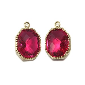 Może przedstawiać: Dwa złote zawieszki z czerwonym, ośmiokątnym kamieniem szklanym w środku. Kamienie mają fasetowany wzór i są otoczone złotą obwódką.