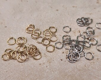May include: A collection of gold and silver jump rings. The jump rings are small and circular, and they are arranged in a pile on a light brown surface.