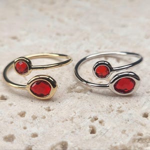 Può includere: Due anelli con design a bypass, ciascuno con due pietre rosse. Un anello è dorato, l'altro argentato. Le pietre sono ovali e rotonde, incastonate in una lunetta. Gli anelli sono esposti su una superficie chiara e strutturata.