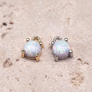 May include: Two small, round opal charms with a silver and gold setting. The charms are on a light brown surface.
