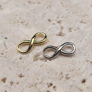 Pode incluir: Dois pingentes com símbolo de infinito, um dourado e outro prateado, sobre uma superfície texturizada bege claro.