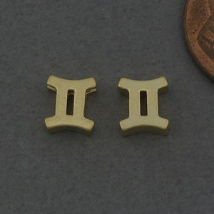 Könnte beinhalten: Zwei goldfarbene Metall-Anhänger in Form des Zwillinge-Sternzeichen-Symbols. Die Anhänger sind etwa so groß wie eine deutsche Ein-Cent-Münze.