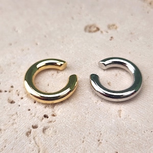 May include: Two ear cuffs, one gold and one silver, are shown on a light beige textured surface. The ear cuffs are simple, round, and open on one side.
