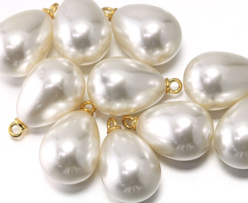 May include: A group of ten white pearlescent teardrop-shaped beads with gold-colored metal loops at the top.