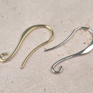May include: Two earring hooks, one gold and one silver, are shown on a light brown surface. The hooks are curved and have a small loop at the end.