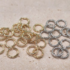May include: A close-up of two groups of small, gold and silver jump rings. The rings are textured and have a rounded shape.