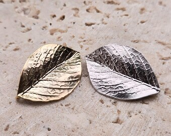 May include: Two leaf-shaped metal ornaments. One is gold-colored, the other is silver. Both have detailed veining and a textured surface. The leaves are displayed on a light-colored, textured surface.