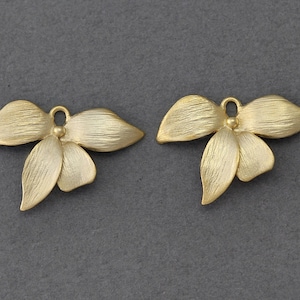 Może przedstawiać: Dwa złote metalowe charms w kształcie kwiatu orchidei. Charms mają fakturowane, szczotkowane wykończenie i są w kształcie orchidei z pięcioma płatkami.