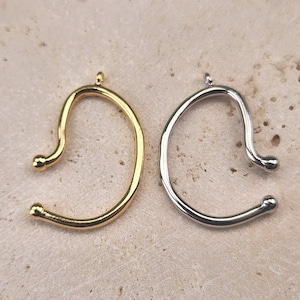 Könnte beinhalten: Zwei Ohrringe, einer golden und einer silbern, werden auf einer hellbraunen, strukturierten Oberfläche gezeigt. Die Ohrringe sind in Form einer geschwungenen Linie mit einer kleinen Kugel an jedem Ende gestaltet.