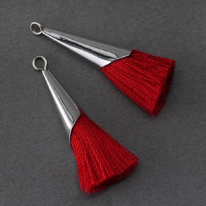 May include: Two red tassel pendants with silver-coloured metal caps and loops. The tassels are made of fine, vibrant red threads. The pendants are displayed on a dark grey surface, showcasing their design and colour.