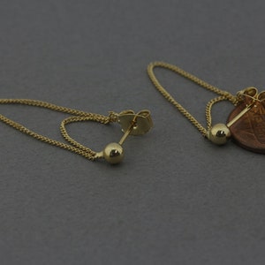 Puede incluir: Un par de pendientes de oro con una cadena que cuelga de cada uno. Un pendiente tiene una pequeña bola de oro al final de la cadena, mientras que el otro pendiente tiene un dije redondo en forma de moneda.