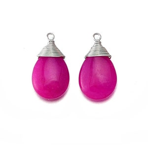 May include: Two magenta teardrop-shaped pendants with silver wire wrapped around the top. The pendants are a vibrant pink color and have a smooth, polished surface. The silver wire forms a conical shape at the top, with a small loop for hanging. The background is white.