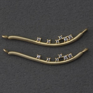 May include: Two gold-toned metal jewelry pieces with a curved shape and small clear gemstones set in the metal. The pieces are similar in shape and size.
