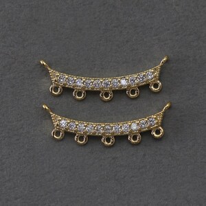 May include: Two gold-colored, curved jewelry components. Each component features a row of clear, sparkling stones and several small loops for attachment. The ends have a decorative, pointed design.