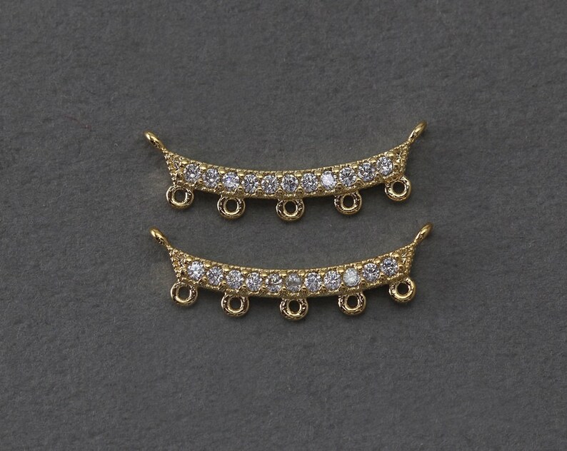 May include: Two gold-colored, curved jewelry components. Each component features a row of clear, sparkling stones and several small loops for attachment. The ends have a decorative, pointed design.