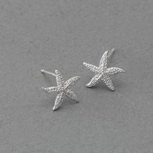 Puede incluir: Un par de pendientes de estrella de mar plateados. Los pendientes son pequeños y delicados, y tienen un acabado brillante.
