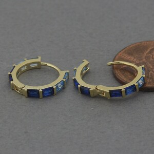 May include: A pair of gold hoop earrings with blue and light blue rectangular gemstones. The earrings are about the size of a US penny.