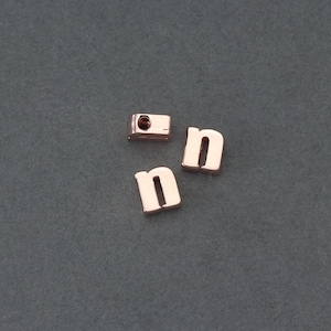 May include: Three gold-colored letter beads, each spelling the letter "n". The beads are on a gray surface.