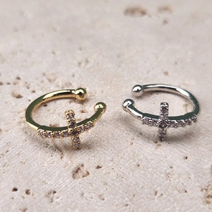 May include: Two ear cuffs with a cross design. The ear cuff on the left is gold with clear rhinestones. The ear cuff on the right is silver with clear rhinestones.