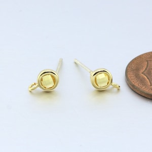 May include: A pair of gold-tone earring studs with a small, clear, faceted gemstone in the center of each stud. The earrings are shown next to a US penny for size comparison.