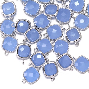 May include: A group of light blue glass square-shaped charms with silver metal accents. The charms have a faceted design and are scattered on a white surface.