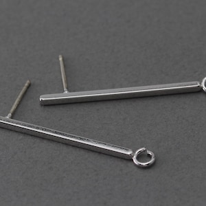 May include: Two silver bar earrings with a post and a small loop at the end. The earrings are simple and modern, with a minimalist design. The earrings are shown on a gray background.