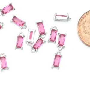 May include: A group of pink crystal rectangle shaped charms with silver jump rings. The charms are scattered on a white surface.