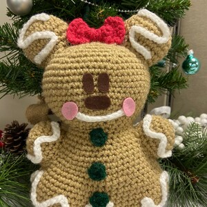 May include: A crocheted gingerbread person toy with a red bow, brown eyes, pink cheeks, and green buttons. The toy is brown with white icing details and is sitting on a white surface.
