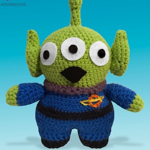 Puede incluir: Un juguete extraterrestre de crochet verde con tres ojos, un pico negro y un mono azul con un cinturón negro y un símbolo espacial amarillo y naranja.