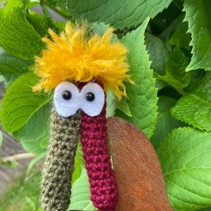 May include: A crocheted worm toy with a burgundy and green body, white eyes, and a yellow fuzzy hairdo. The toy is sitting on a rusty metal surface.