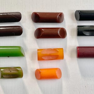May include: A collection of ten cylindrical, vintage drawer pulls in various colors, including brown, green, orange, and black. Each pull has rounded ends and a smooth, glossy finish. The pulls are arranged on a white surface.