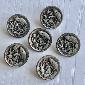 May include: Six silver-toned buttons with a raised design of two birds in a wreath of leaves. The buttons are round and have a slightly raised edge.