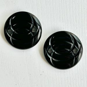 May include: Two round, black buttons with a glossy finish. Each button features a carved design of intersecting lines and shapes, creating a geometric pattern. The buttons are likely for sewing or crafting projects.