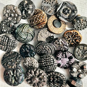 May include: A collection of vintage buttons in various shapes, sizes, and colors. The buttons feature intricate designs, including floral patterns, geometric shapes, and textured surfaces. Colors range from silver and gold to black and iridescent hues.