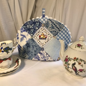 May include: A blue and white patchwork tea cozy with a scalloped edge. The cozy is decorated with a small teapot design and buttons. A white teapot with a floral design is in the background.