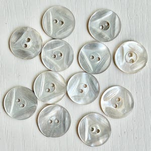 May include: A collection of twelve circular, off-white buttons with a pearlescent finish. Each button features a smooth, slightly concave surface with three distinct, raised diagonal ridges and two central holes for attachment.