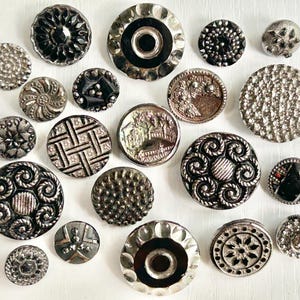 May include: A collection of twenty assorted round buttons in various sizes, featuring metallic silver and black finishes. The buttons display diverse textures, including intricate floral patterns, geometric grids, swirling scrolls, faceted surfaces, and smooth centers.