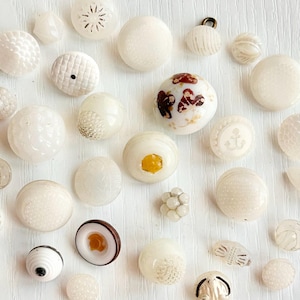 May include: A collection of vintage white buttons in various shapes and textures. Some buttons are smooth, others have intricate patterns, and one features a floral design. The buttons are scattered across a light surface, showcasing their diverse designs.