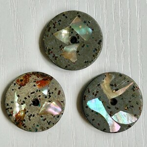 May include: Three circular buttons featuring a dark gray base with embedded iridescent shell fragments and small metallic flecks. Each button has a single central hole for attachment, showcasing a textured surface with varied light-reflecting pieces.
