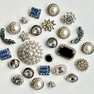 May include: A collection of decorative buttons in various shapes and sizes. The buttons feature pearls, rhinestones, and crystals in silver, blue, and black. Some buttons have a pearl center, while others have a clear or black center.