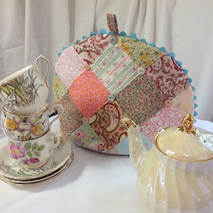 May include: A patchwork tea cozy with a blue trim and buttons, a teapot, and three teacups and saucers with floral designs.