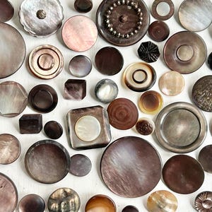 May include: A collection of various shaped buttons in shades of brown, gray, and iridescent colors. The buttons include round, square, and geometric shapes, with some featuring decorative details. The buttons are arranged on a white surface.