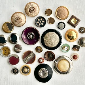 May include: A collection of vintage buttons in various sizes, shapes, and materials. The buttons include gold, silver, black, and brown tones, with textures like fabric, metal, and plastic. Some buttons have intricate designs and patterns.
